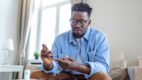man is sitting at the sofa and taking blood from his finger due to diabetes. The daily life of a man of African-American ethnicity person with a chronic illness who is using glucose tester.